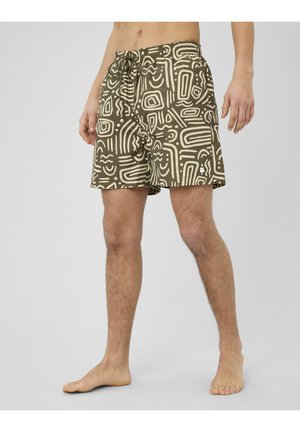 Homme portant un short de bain vert olive avec des motifs géométriques abstraits blancs, debout pieds nus sur un fond clair uni.