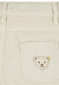 Pantaloni in cotone beige chiaro con una tasca posteriore decorata con un ricamo a forma di faccia di orsetto marrone e bianco, e un piccolo dettaglio giallo.