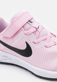 Scarpa sportiva rosa chiaro con tomaia in rete, logo Nike Swoosh nero e cinturino regolabile. Presenta una superficie testurizzata e una suola intermedia bianca.