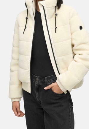 Person trägt eine cremefarbene Steppjacke mit hohem Kragen und schwarzem Reißverschluss, kombiniert mit schwarzen Jeans und einem schwarzen Shirt, die Hand in der Tasche.