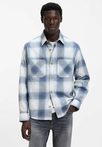 Blauw-wit geruit overhemd van zachte stof met lange mouwen, knoopsluiting aan de voorkant en twee borstzakken. Gedragen over een wit T-shirt.