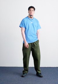 Zils īsām piedurknēm krekls ar Nike logo, apvienots ar brīvām zaļām biksēm un melniem sporta apaviem. Stāv uz pelēka grīdas pretī baltām sienām.