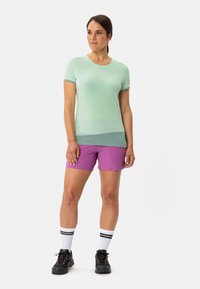 Light green short-sleeve athletic shirt with contrast trim, paired with purple shorts. Model wears black hiking shoes and white socks with black stripes.