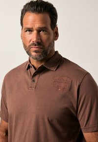 Hnědá polokošile vyrobená z bavlny, s texturovanou nášivkou loga na hrudi, dvoutlačítkovou légou a krátkými rukávy.