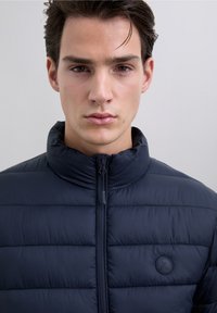 Granatowa kurtka puchowa z wysokim kołnierzem, zapięciem na zamek i okrągłą naszywką z logo. Powierzchnia jest gładka, z pikowanym wzorem dla lepszej izolacji.