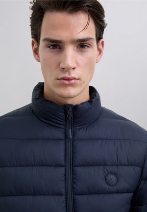 Chaqueta de plumas azul marino con cuello alto, cierre de cremallera y parche de logo redondeado. La superficie es lisa y cuenta con un diseño acolchado para aislamiento.