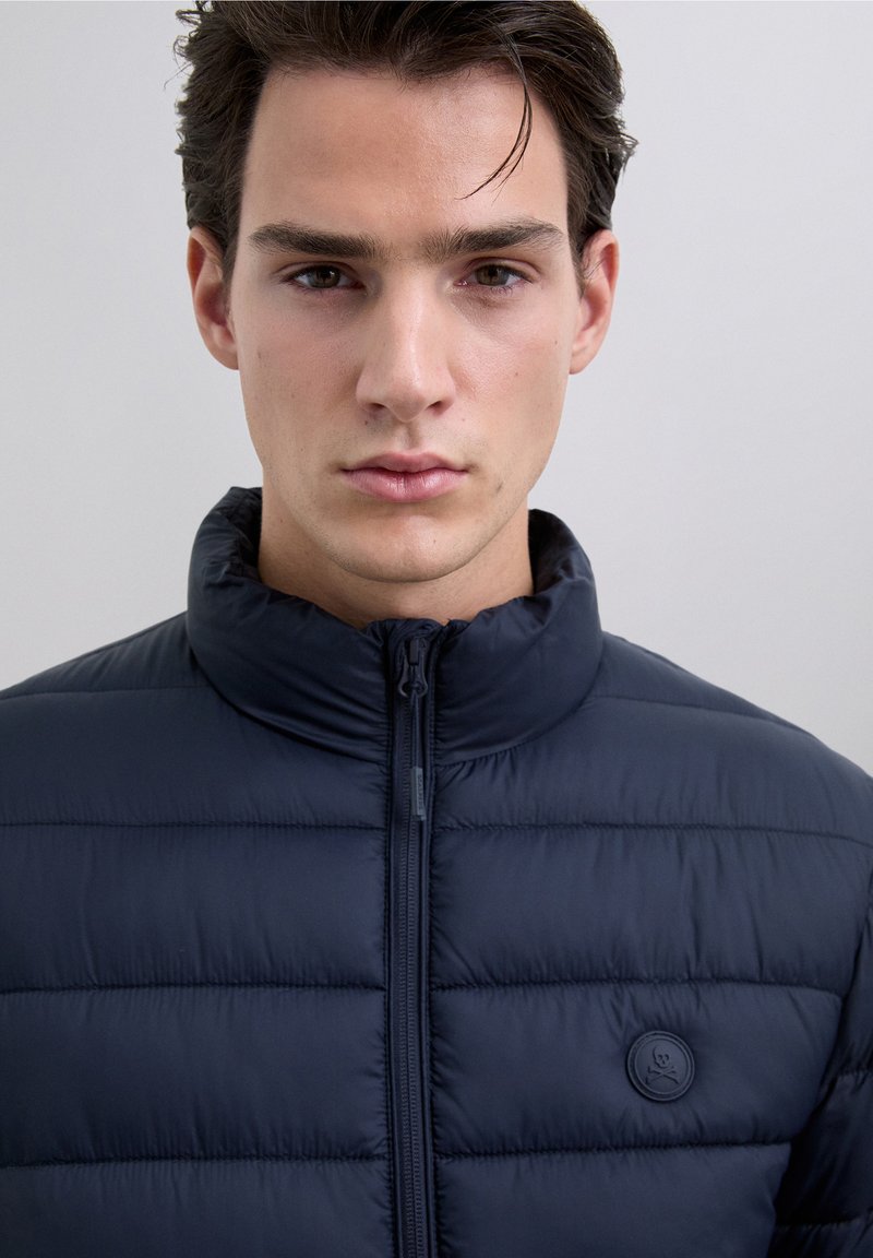 Granatowa kurtka puchowa z wysokim kołnierzem, zapięciem na zamek i okrągłą naszywką z logo. Powierzchnia jest gładka, z pikowanym wzorem dla lepszej izolacji.