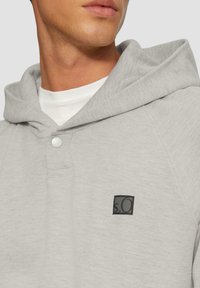Sudadera gris hecha de un tejido suave con una tapeta de botones y un parche de logo negro. Cuenta con una capucha grande y un cuello de canalé.