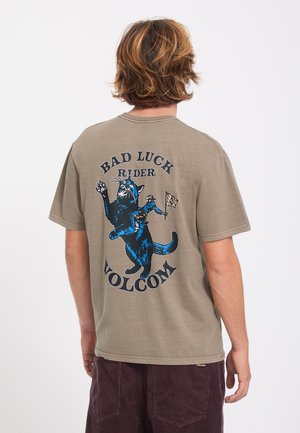 Personne aux cheveux châtain clair portant un t-shirt marron avec un chat bleu chevauchant un plus petit chat et le texte "Bad Luck Rider Volcom" dans le dos.