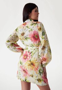 Kwiatowa sukienka z lekkiego materiału, z żywymi różowymi i żółtymi kwiatami, długimi rękawami i paskiem w talii, który podkreśla sylwetkę.