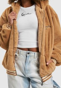 Kurtka z beżowego futerka sztucznego z zamkiem z przodu i ściągaczami na mankietach, noszona na białej koszulce z czarnym haftowanym napisem, w połączeniu z jasnoniebieskimi szortami jeansowymi.