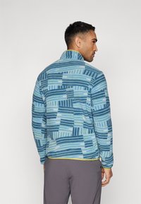 Fleece pulover u plavim tonovima, s geometrijskim uzorcima i visokim ovratnikom. Uključuje žute akcentne detalje i duge rukave, s opuštenim krojem.