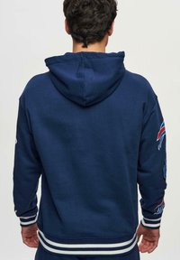 Marinblå huvtröja med vita randiga muddar och nederkant, prydd med färgglada tryck på ärmarna, tillverkad av mjukt tyg, bakvy.