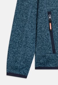 Fleecejakke i mørk blå med teksturert overflate, utstyrt med en glidelåslomme med oransje detaljer og ribbestrikkede mansjetter.