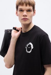 Μαύρο βαμβακερό μπλουζάκι με λευκό γραφικό σχέδιο που περιλαμβάνει τη λέξη "HUGO" μέσα σε κυκλικό τύπωμα, με κοντά μανίκια και κλασικό στρογγυλό λαιμό.