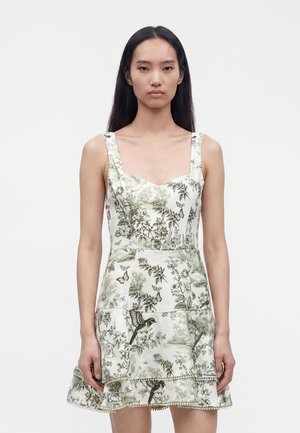 Jeune femme aux longs cheveux foncés porte une robe blanche sans manches avec des motifs floraux et d'oiseaux verts sur un fond clair uni.
