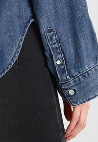 Manche de chemise en denim avec poignet boutonné reposant à côté d'une main, associée à un pantalon noir sur un fond neutre.