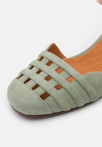Chaussure plate en daim vert clair avec bout fermé et design à bride tressée, avec une semelle intérieure rembourrée couleur camel et une semelle extérieure marron.