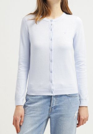 Femme portant un cardigan bleu clair boutonné et un jean bleu clair avec une petite déchirure sur la cuisse droite.
