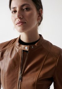 Chaqueta de cuero marrón con cuello alzado, cremallera plateada y botones a presión. Textura suave con costuras ajustadas y diseño minimalista.