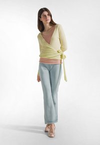 Joven mujer con un suéter cruzado amarillo, top rosa, pantalones azul claro y zapatos de ballet rosas, de pie contra un fondo blanco.