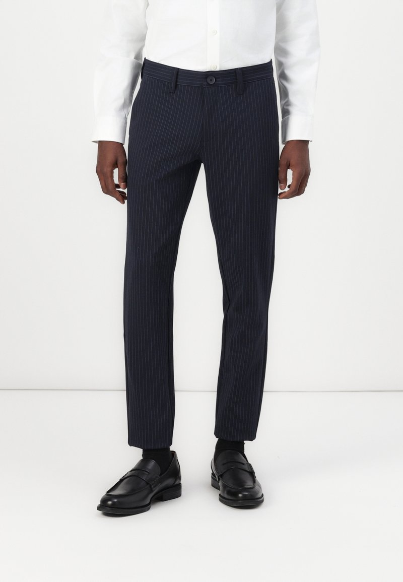 Homme portant un pantalon de costume bleu marine à rayures, une chemise blanche, des chaussettes noires et des mocassins en cuir noir, debout devant un fond blanc.