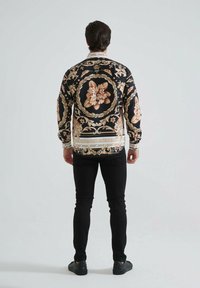 Chemise noire à manches longues ornée de motifs floraux crème et rose, avec une bordure dorée décorative et un col blanc contrastant, portée avec un pantalon noir.