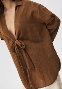 Femme portant une chemise en tissu texturé brun ample nouée à l'avant, avec les manches retroussées et un pantalon de couleur claire visible en dessous.