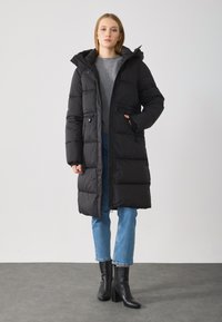 Cappotto imbottito nero con cappuccio rimovibile, chiusura con zip e due tasche anteriori. Abbinato a jeans blu chiaro e stivaletti neri con tacco.