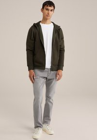 Haut à capuche zippé vert avec poches avant, superposé sur un t-shirt blanc, associé à un jean gris clair et des baskets blanches.