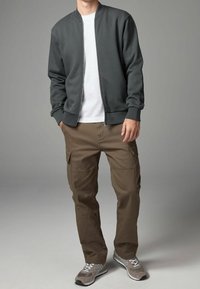 Veste bomber gris foncé portée sur un t-shirt blanc, associée à un pantalon cargo marron et des chaussures de sport grises, avec plusieurs poches.
