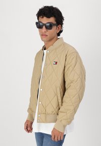 Chaqueta bomber acolchada beige con botones de presión plateados, cuello de canalé y parche con logo. Llevada sobre una camisa blanca y combinada con jeans azules.