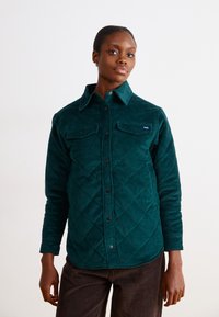 Chemise en velours côtelé vert avec un design matelassé, fermeture à boutons sur le devant et deux poches poitrine. Dotée d'un col et de manches longues.