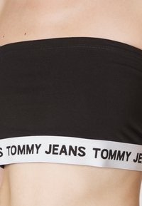 Czarny strapless crop top wykonany z gładkiego materiału, z białą elastyczną taśmą z nadrukiem "TOMMY JEANS" w czerni.