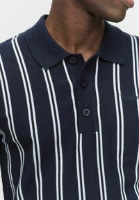 Marinblå polo skjorta med vita vertikala ränder, strukturerad kvalitet, två svarta knappar och kragadetalj, med varumärkesnamnet broderat på sidan.