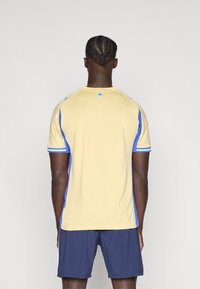 Κίτρινο αθλητικό μπλουζάκι με μπλε λεπτομέρειες και κοντά μανίκια, με υφή στο ύψος των ώμων και λογότυπο κοντά στη γραμμή του λαιμού.