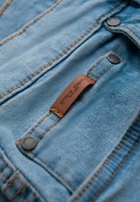 Tela de denim azul con una superficie ligeramente texturizada, que presenta una etiqueta de cuero marrón y botones metálicos a lo largo de la costura.