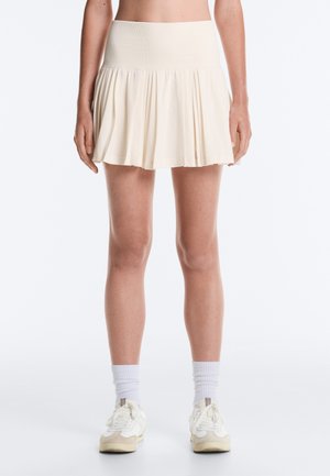 Mini-jupe plissée crème portée par une personne debout, associée à des chaussettes blanches et des baskets blanches sur un fond uni.