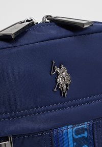 Laivastonsininen nailonlaukku, jossa on hopeinen logon tunnus, kiiltävät metallivetoketjut ja sininen kuvio sivulla, teksturoidulla ompelu-yksityiskohdalla.