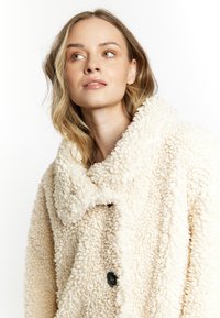 Krémový huňatý kabát vyrobený z texturované tkaniny, s širokým límcem a černými knoflíky. Design je oversized a pohodlný.