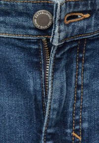 Denim-farkut, joissa on tumma sininen pesu, vetoketju ja metallinen nappi, jossa on leima "ESTABLISHED 1984", ja kontrastiväriset ruskeat ompeleet.