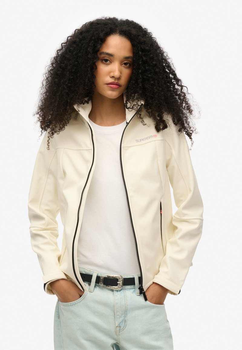 Crème-kleurige softshell jas met hoge kraag, volledige ritssluiting en zwarte accessoires. Voorzien van zijzakken en subtiele branding.