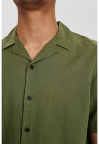 Camicia verde a maniche corte con colletto, bottoni neri e un tessuto testurizzato, mostrata parzialmente dalla zona del collo in giù.