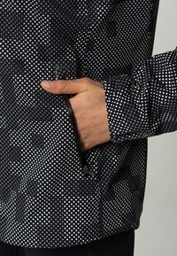 Veste noire arborant un motif géométrique de petits carrés blancs, avec une poche zippée sur le côté droit et une texture lisse et résistante à l'eau.