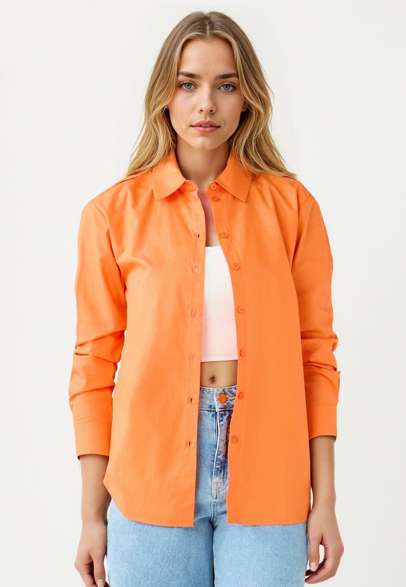 Camicia arancione con colletto, maniche lunghe e vestibilità rilassata. Realizzata in tessuto morbido. Indossata sopra un crop top bianco e jeans di denim.