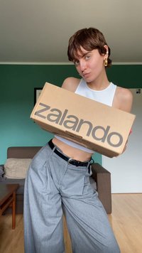 Zalando kartoninė dėžė su didžiuliais pilkais užrašais, kurią laiko žmogus, vilkintis baltą marškinėlį be rankovių ir pilkas plisuotas kelnes, atsukęs kamerai.