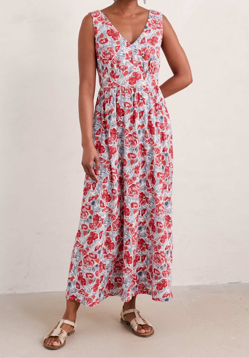 Femme portant une robe maxi sans manches à encolure en V avec un motif floral rouge et rose sur un fond bleu clair, associée à des sandales beige.