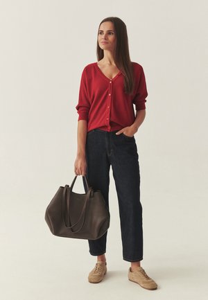 Sieviete, tērpusies sarkanā piepogājamā kardiganā, tumšās džinsās un bēšās sporta kurpēs, tur lielu tumši brūnu rokassomu, stāvot pret gaišu fonu.