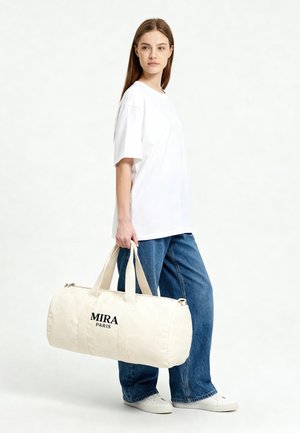 Femme portant un t-shirt blanc et un jean, tenant un grand sac de sport couleur crème avec l'inscription "MIRA PARIS", debout devant un fond uni.