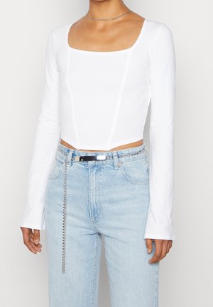 Vrouw die een witte, nauwsluitende, lange mouwen, vierkante hals top draagt die is ingestopt in lichtblauwe jeans met een hoge taille en een zilveren kettingriem.